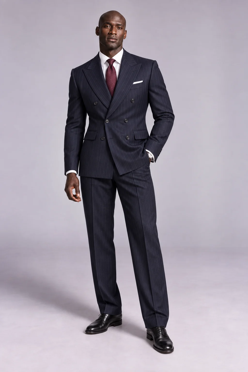 A tall bald dark-skinned man wearing a fitted navy pinstripe double-breasted suit, white shirt, burgundy tie and black Oxford shoes